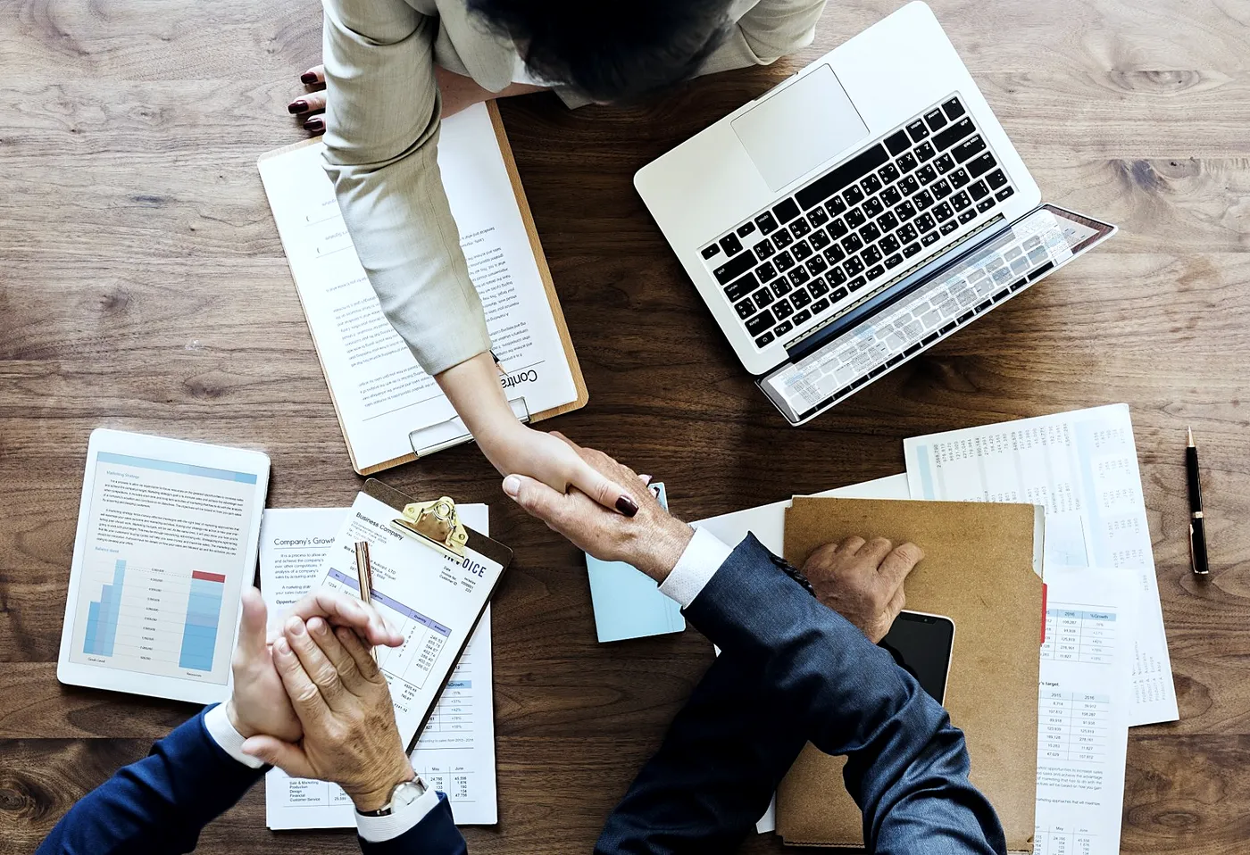 Duas pessoas apertando as mãos sobre uma mesa de madeira com contrato, notebook e relatórios