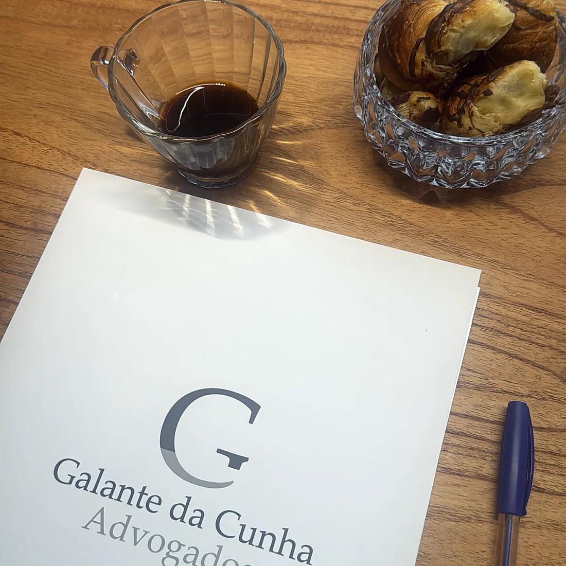 Café servido sobre a mesa de madeira, ao lado de papel com a marca do escritório e uma caneta