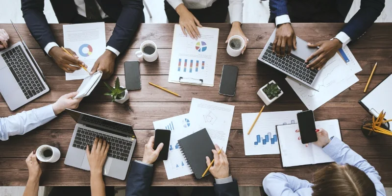 Equipe reunida em volta de uma mesa de madeira, com relatórios, notebooks e café